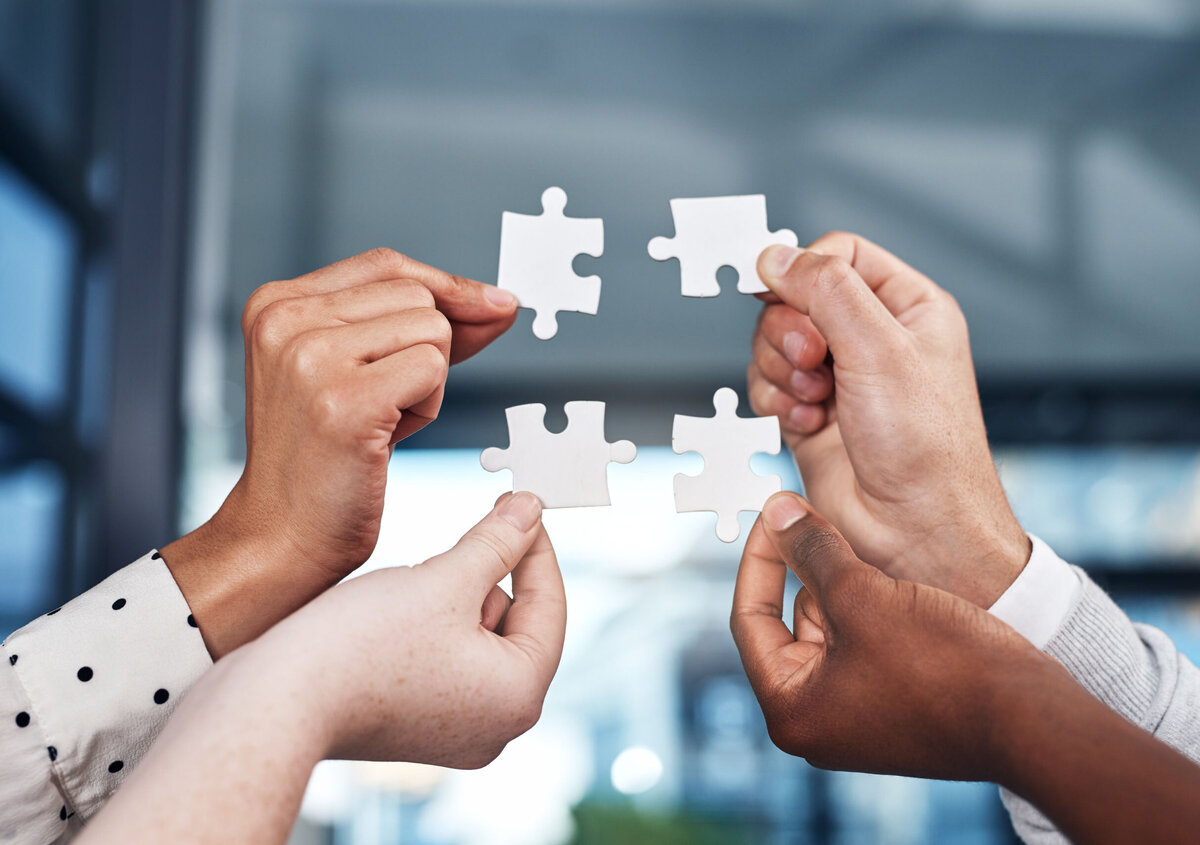 Mehrere H&auml;nde halten wei&szlig;e Puzzleteile und f&uuml;hren diese zusammen. Das Bild symbolisiert Zusammenarbeit, Partnerschaft und gemeinsame Probleml&ouml;sung.