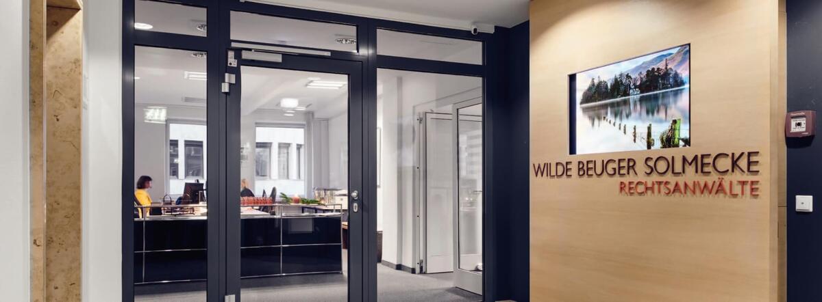 Entrance area of the Cologne-based law firm WILDE BEUGER SOLMECKE, featuring a modern glass-fronted reception area. The firm's name, &laquo;WILDE BEUGER SOLMECKE RECHTSANW&Auml;LTE,&raquo; is displayed on a wood-paneled wall, with office spaces visible through the glass doors in the background.
