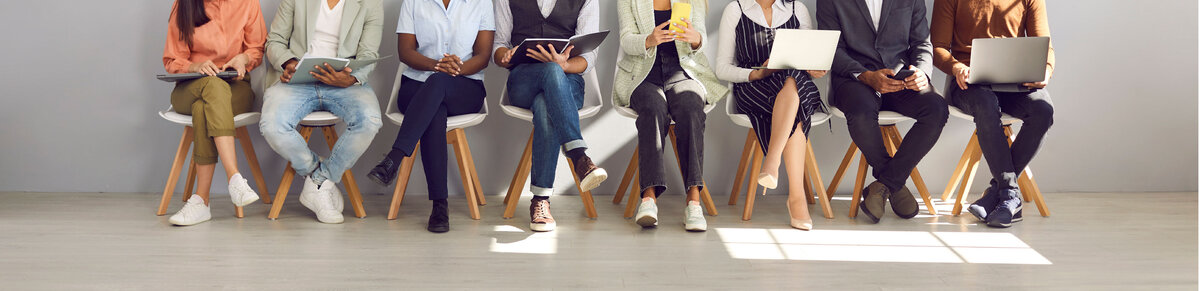 Meerdere sollicitanten zitten naast elkaar met laptops, tablets en smartphones terwijl zij wachten op een sollicitatiegesprek. De afbeelding symboliseert carri&egrave;remogelijkheden, werving, talentontwikkeling en professionele groei.