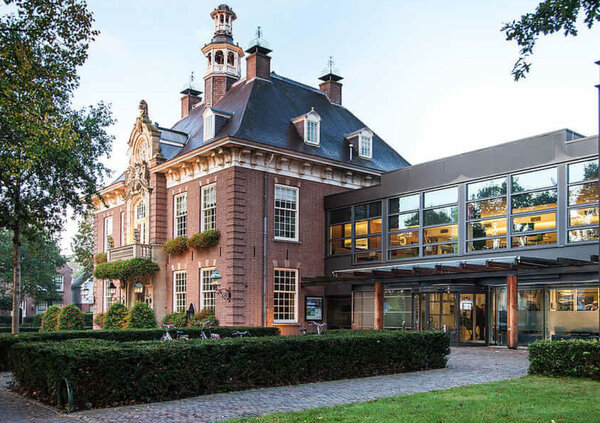 Historisk tegelbyggnad med karakt&auml;rsfull arkitektur, ett brant mansardtak och ett litet torn. Till h&ouml;ger &auml;r en modern tillbyggnad ansluten, med en stor glasfasad och en inglasad entr&eacute;. V&auml;lsk&ouml;tta h&auml;ckar, gr&auml;smattor och en stenlagd g&aring;ngv&auml;g leder fram till ing&aring;ngen. Flera cyklar st&aring;r parkerade framf&ouml;r byggnaden. Fotograferad i dagsljus framh&auml;ver bilden den lyckade kombinationen av historisk och modern arkitektur.
