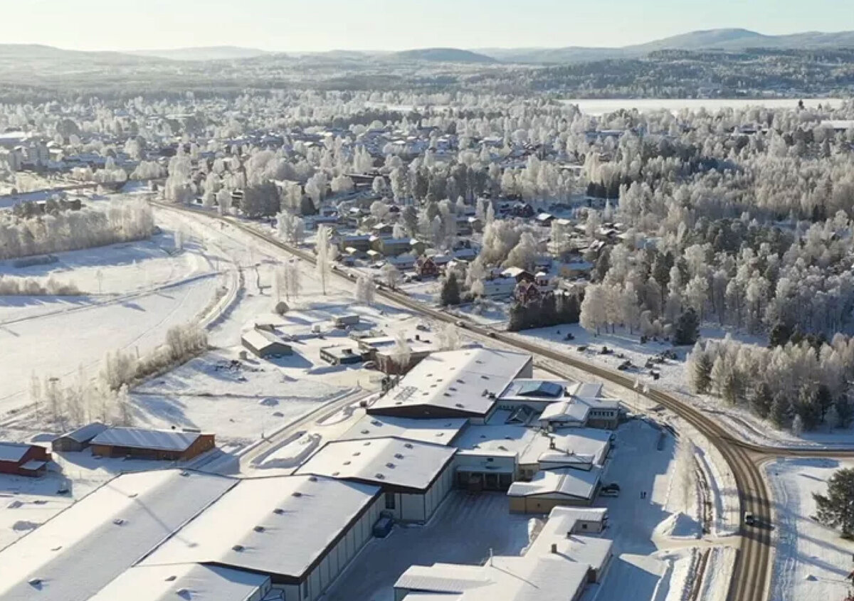 Luchtfoto van een besneeuwd winterlandschap. Op de voorgrond bevindt zich een groot industri&euml;le- en kantorencomplex, bestaande uit meerdere magazijn- en kantoorgebouwen met met sneeuw bedekte daken. De omliggende wegen, parkeerterreinen en open ruimtes zijn eveneens bedekt met een laag sneeuw. De afbeelding benadrukt de omvang van het terrein en de ligging binnen een uitgestrekt winterlandschap.