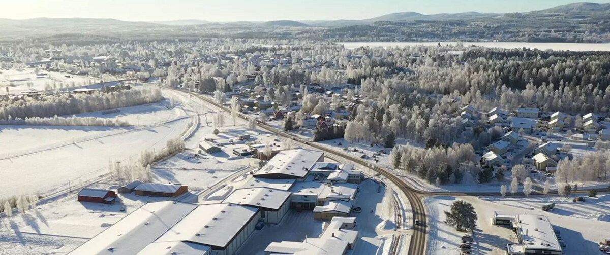 Luchtfoto van een besneeuwd winterlandschap. Op de voorgrond bevindt zich een groot industri&euml;le- en kantorencomplex, bestaande uit meerdere magazijn- en kantoorgebouwen met met sneeuw bedekte daken. De omliggende wegen, parkeerterreinen en open ruimtes zijn eveneens bedekt met een laag sneeuw. De afbeelding benadrukt de omvang van het terrein en de ligging binnen een uitgestrekt winterlandschap.