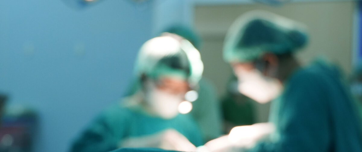 Vervaagde opname van een chirurgische ingreep in een operatiekamer, waarbij medische professionals in beschermende kleding rond een pati&euml;nt werken onder felle operatielampen.