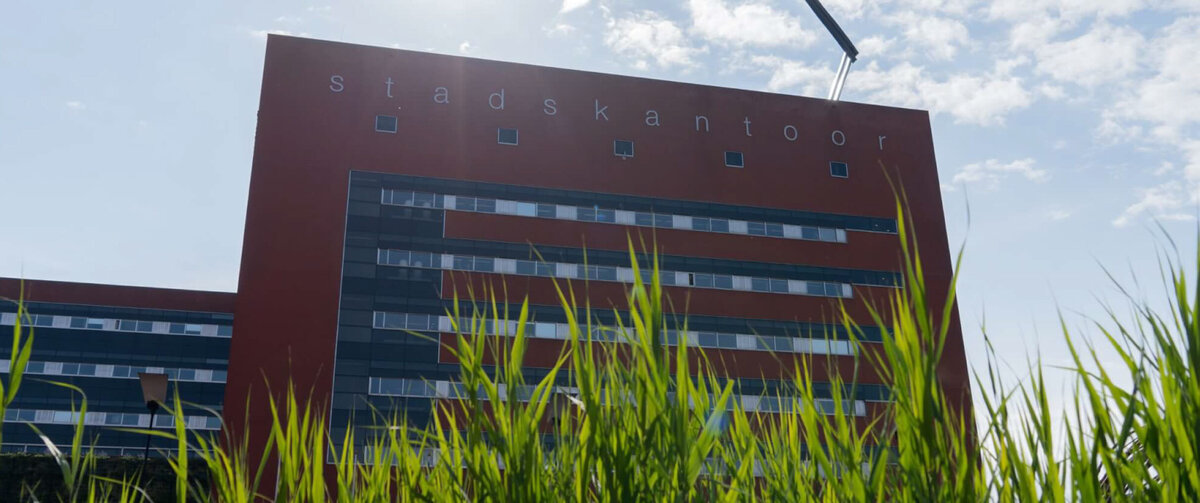 Modern kontorsbyggnad f&ouml;r Milj&ouml;myndigheten IJsselland med en r&ouml;d fasad och texten &raquo;Stadskantoor&raquo;, fotograferad ur l&aring;g vinkel bakom h&ouml;gt gr&ouml;nt gr&auml;s under en solig himmel.