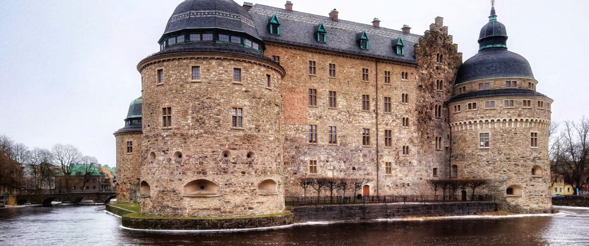 Historisch kasteel &Ouml;rebro in Zweden, een grote stenen vesting met ronde torens en donkere koepeldaken, omgeven door water onder een bewolkte hemel.