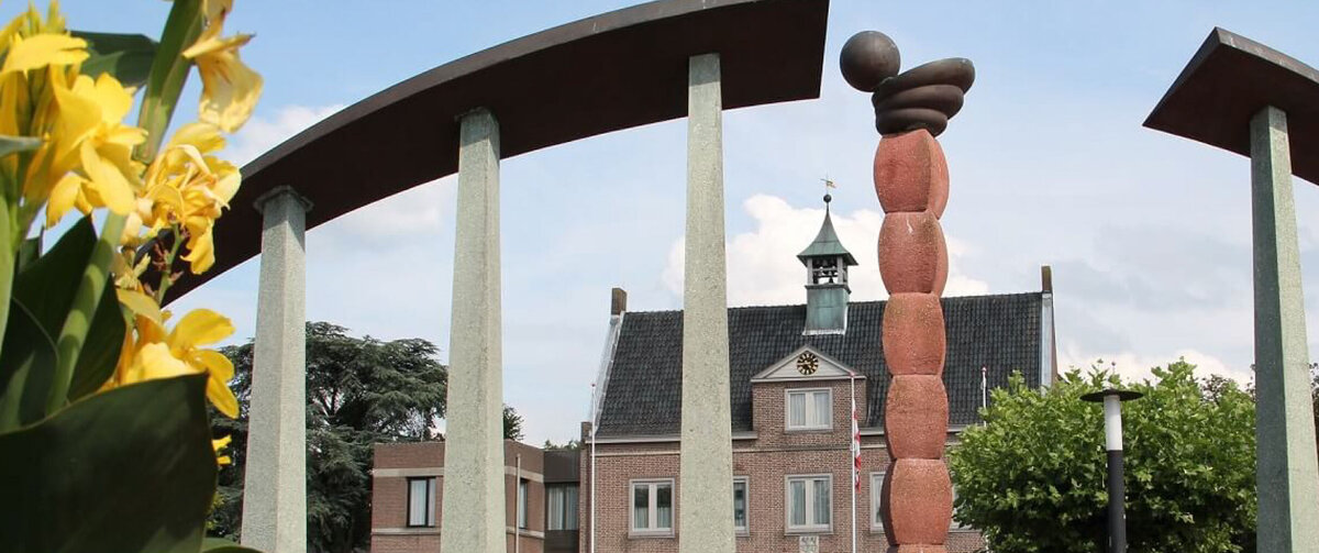 R&aring;dhustorget i Someren i Nederl&auml;nderna, med en modern offentlig konstskulptur inramad av b&ouml;jda stenpelare. F&auml;rgglada blommor pryder f&ouml;rgrunden, medan kommunhuset och tr&auml;d syns i bakgrunden under en klarbl&aring; himmel.