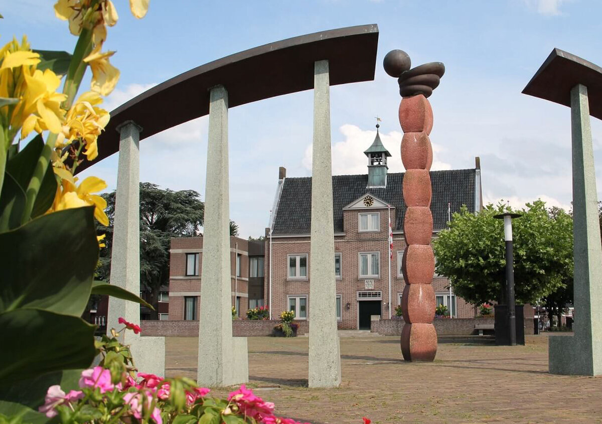 Dorpsplein in Someren, Nederland, met een modern kunstwerk, omlijst door gebogen stenen zuilen. Op de voorgrond bloeien kleurrijke bloemen, terwijl het gemeentehuis en bomen zichtbaar zijn op de achtergrond onder een heldere blauwe hemel.