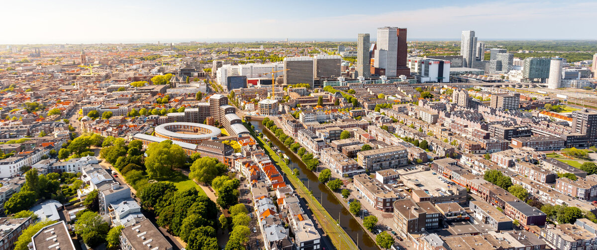 Flygvy &ouml;ver ett vidstr&auml;ckt stadslandskap en solig dag. I f&ouml;rgrunden syns bostadsbyggnader, tr&auml;dkantade gator och en rak kanal som l&ouml;per genom omr&aring;det. I mitten av bilden breder t&auml;ttbebyggda stadsdelar ut sig, med ett karakteristiskt cirkelformat byggnadskomplex som blickf&aring;ng. I bakgrunden reser sig en modern stadssiluett med flera h&ouml;ghus och kontorsbyggnader. Ovanf&ouml;r staden str&auml;cker sig en klarbl&aring; himmel med endast n&aring;gra f&aring; moln.