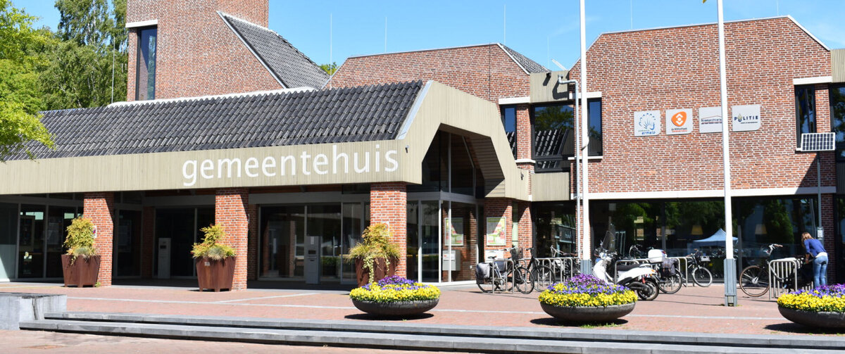 Front view of the Ermelo municipal building in the Netherlands, featuring a brick fa&ccedil;ade, a clock tower, and the entrance marked &laquo;gemeentehuis.&raquo; Bicycles, flower planters, and flagpoles are visible in front of the building under a clear blue sky.