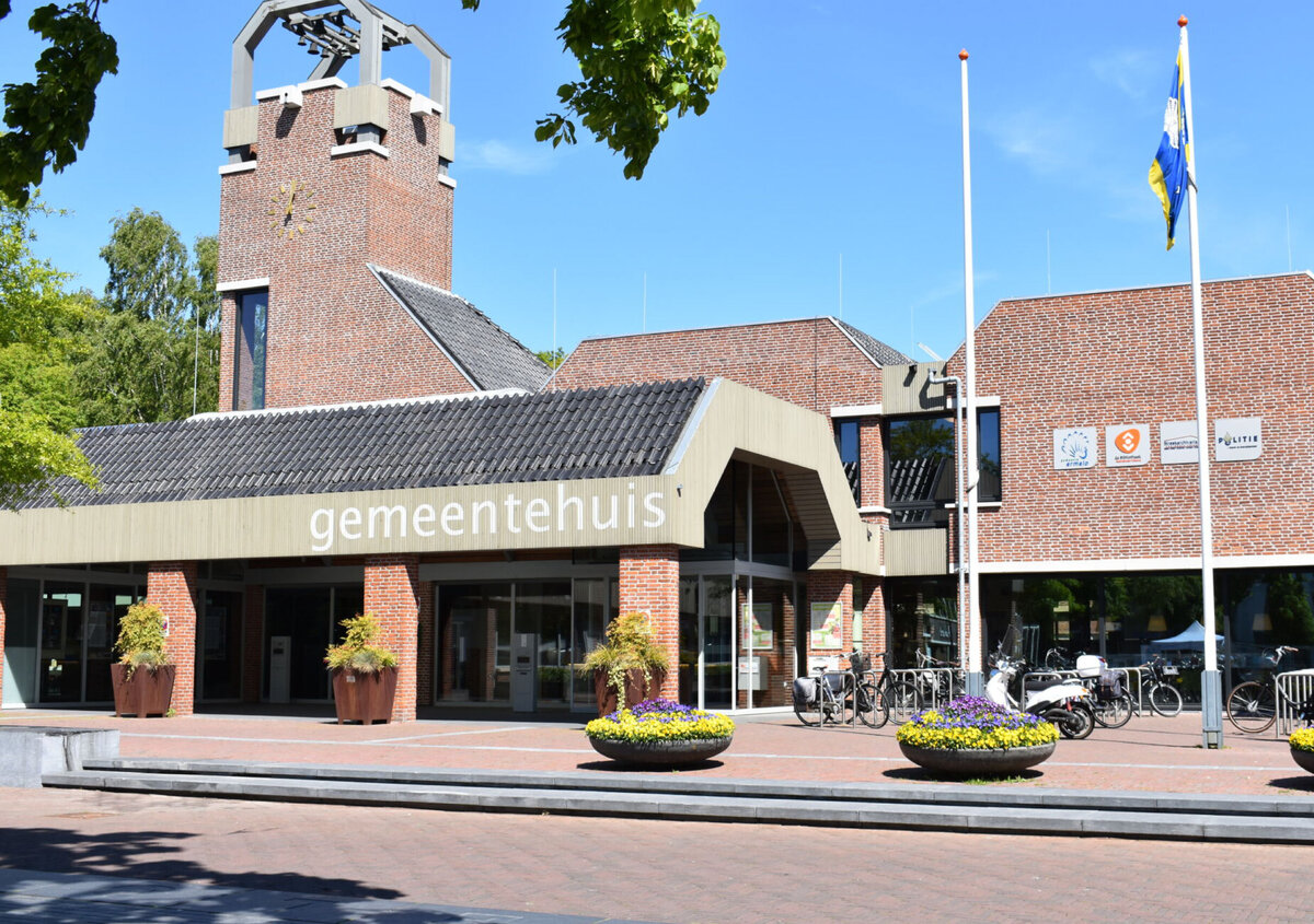 Frontvy av kommunhuset i Ermelo i Nederl&auml;nderna, med en tegelfasad, ett klocktorn och en entr&eacute; m&auml;rkt &raquo;gemeentehuis&raquo;. Framf&ouml;r byggnaden syns cyklar, blomsterplanteringar och flaggst&auml;nger under en klarbl&aring; himmel.