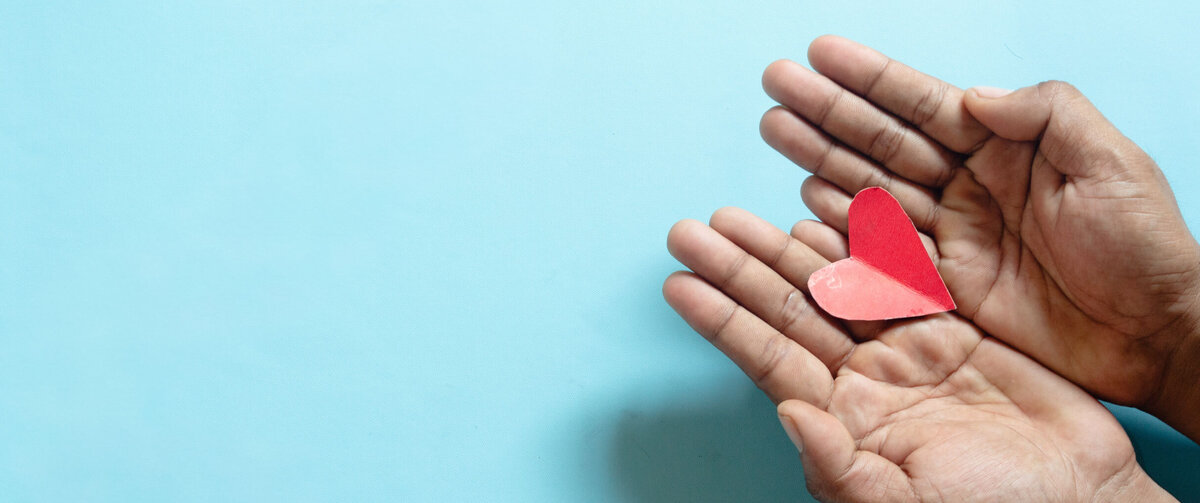 Twee open handen houden voorzichtig een klein rood papieren hart vast tegen een lichtblauwe achtergrond. Het hart ligt centraal in de handpalmen en vormt het duidelijke middelpunt van de afbeelding. De eenvoudige compositie laat veel vrije ruimte rondom het onderwerp, terwijl de zachte kleuren thema's als zorg, ondersteuning, betrokkenheid en maatschappelijke verantwoordelijkheid uitstralen.