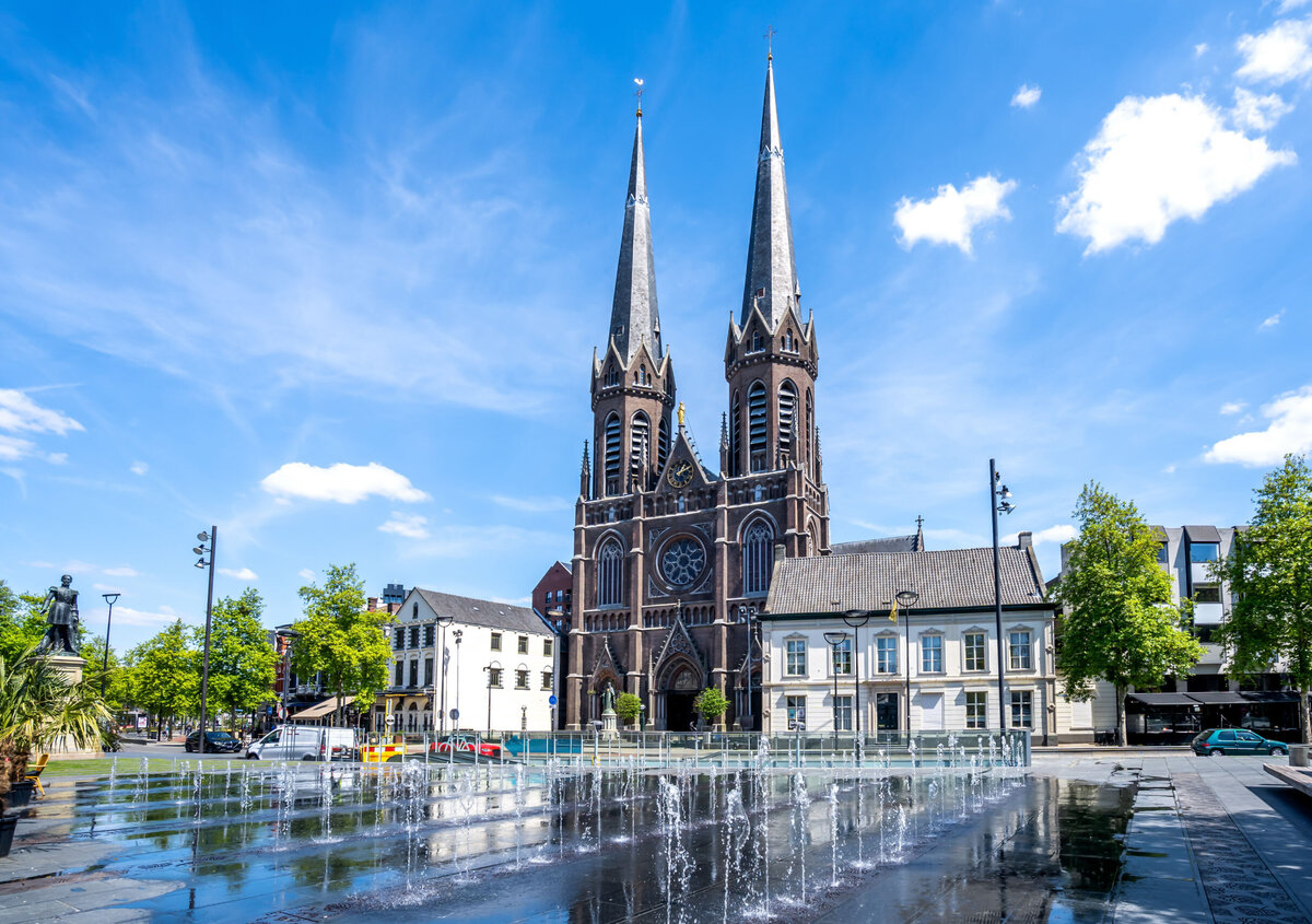Kyrka med tv&aring; spiror som blickar ut &ouml;ver ett stadstorg med font&auml;ner, historiska byggnader och tr&auml;d under en klarbl&aring; himmel.