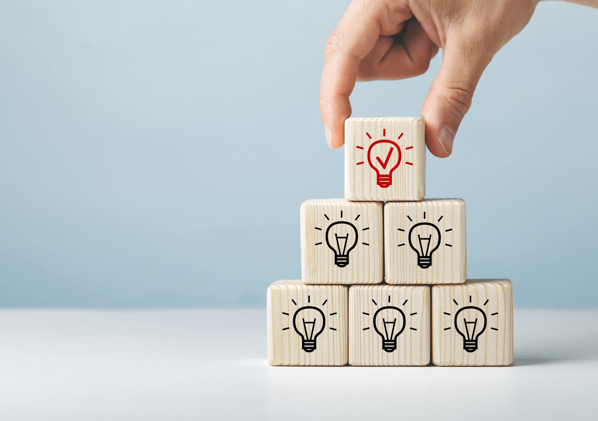 Hand som placerar ett tr&auml;block med en markerad gl&ouml;dlampesymbol h&ouml;gst upp p&aring; en pyramid av tr&auml;block, vilket symboliserar innovation, id&eacute;er, aff&auml;rsl&ouml;sningar och uppt&auml;ckten av nya m&ouml;jligheter genom en SmartDocuments-demo.