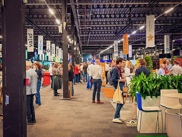 Bild vergr&ouml;&szlig;ern: Drukke beursvloer tijdens CorporatiePlein 2026 met bezoekers die gesprekken voeren bij verschillende stands. Langs de gangpaden hangen banners van exposanten, terwijl in het midden en op de voorgrond meerdere stands, zitgelegenheden en grote plantenbakken zichtbaar zijn. De moderne beurslocatie heeft een industri&euml;le uitstraling met hoge plafonds, donkere draagconstructies en heldere verlichting.