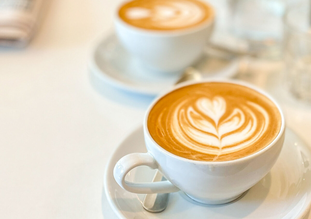 Tv&aring; koppar cappuccino med hj&auml;rtformad latte art p&aring; ett ljust bord. Koppen i f&ouml;rgrunden &auml;r i skarpt fokus, medan den andra koppen och bakgrunden &auml;r mjukt suddiga. Bredvid ligger en tidning och ett glas vatten.