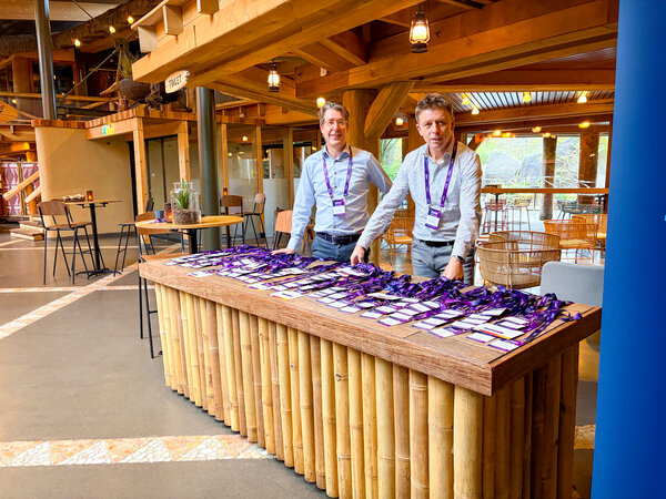 Afbeelding vergroten: Voorbereiding van de registratie voor de SmartDocuments Klantendag 2026. Op een grote ontvangstbalie liggen naamkaartjes en lanyards klaar voor de deelnemers. Op de achtergrond zijn de evenementruimtes zichtbaar in een open, natuurlijk ingerichte locatie.