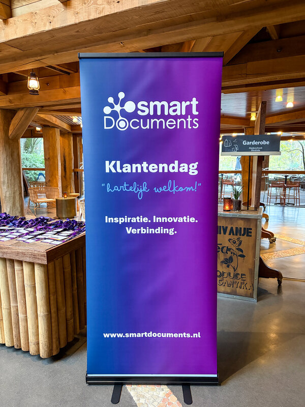 Afbeelding vergroten: SmartDocuments roll-upbanner bij de entree van de Klantendag 2026. Op de banner staan de welkomstboodschap 'Hartelijk welkom!' en de kernbegrippen inspiratie, innovatie en verbinding. Op de achtergrond zijn de registratietafel en de evenementruimtes zichtbaar.