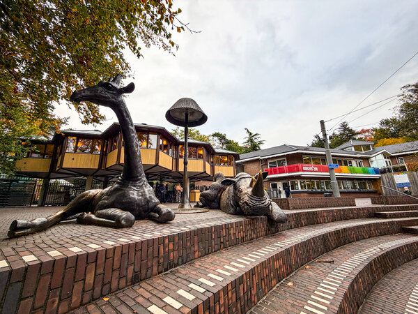 Afbeelding vergroten: Buitenaanzicht van de evenementlocatie in Burgers' Zoo. Op de voorgrond staan grote dierenbeelden van een giraffe en een neushoorn op een terrasvormig plein. Op de achtergrond zijn de gebouwen van de dierentuin en de entree van het evenementencentrum zichtbaar.